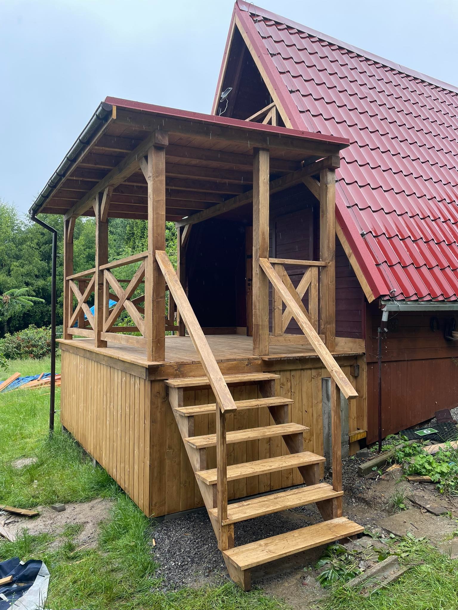 Świeżo wybudowana, drewniana weranda z zadaszeniem, schodami i ozdobnymi balustradami w kształcie litery X, przylegająca do drewnianego domu z czerwonym dachem, widok z zewnątrz w pochmurny dzień.