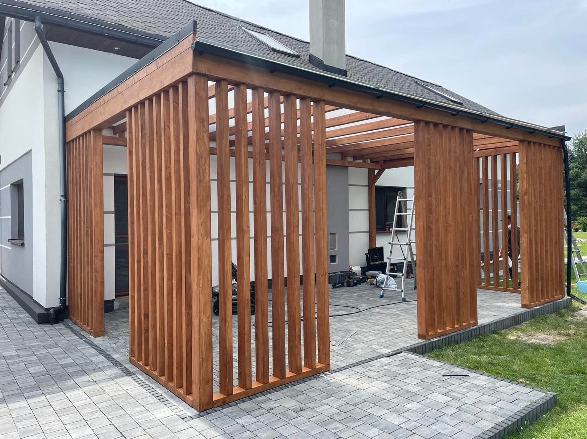 Drewniana pergola z pionowymi listwami, przylegająca do nowoczesnego domu z szarą elewacją, widoczna drabina i narzędzia sugerujące etap montażu.