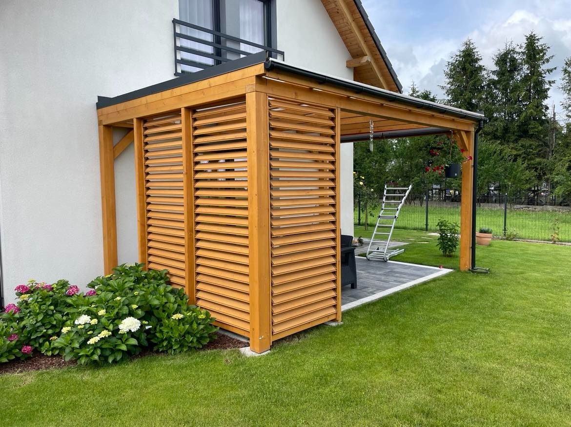 Drewniana pergola z lamelami na tarasie wyłożonym szarą kostką, z drabinką opartą o konstrukcję, obok domu z balkonem i zadbanym trawnikiem.