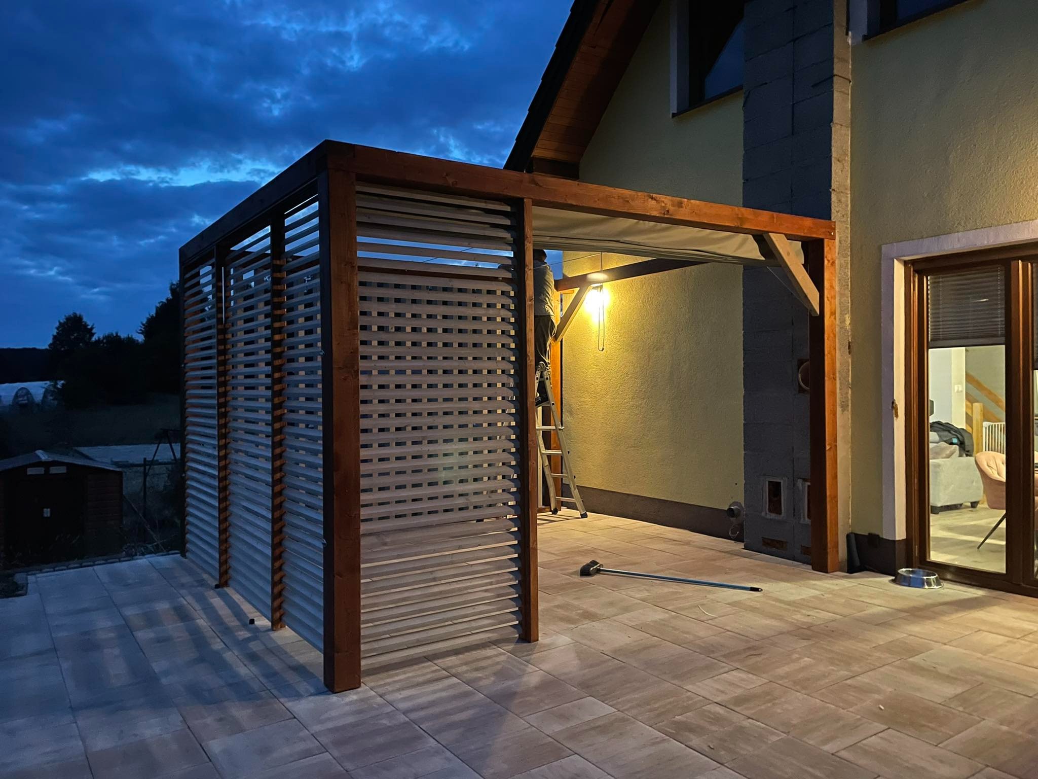 Drewniana pergola z ażurowymi ścianami i zadaszeniem, oświetlona lampą, przylegająca do budynku z widocznym tarasem z kostki brukowej i akcesoriami do czyszczenia.