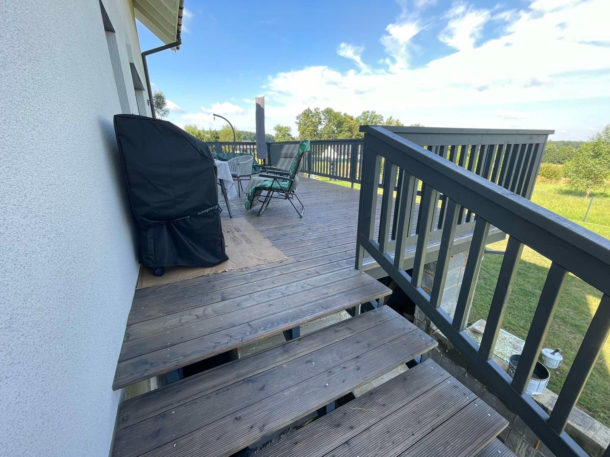 Schody i fragment tarasu z desek w odcieniach szarości, z widoczną osłoną grilla przy ścianie domu i meblami ogrodowymi w tle, uchwycone w słoneczny dzień.