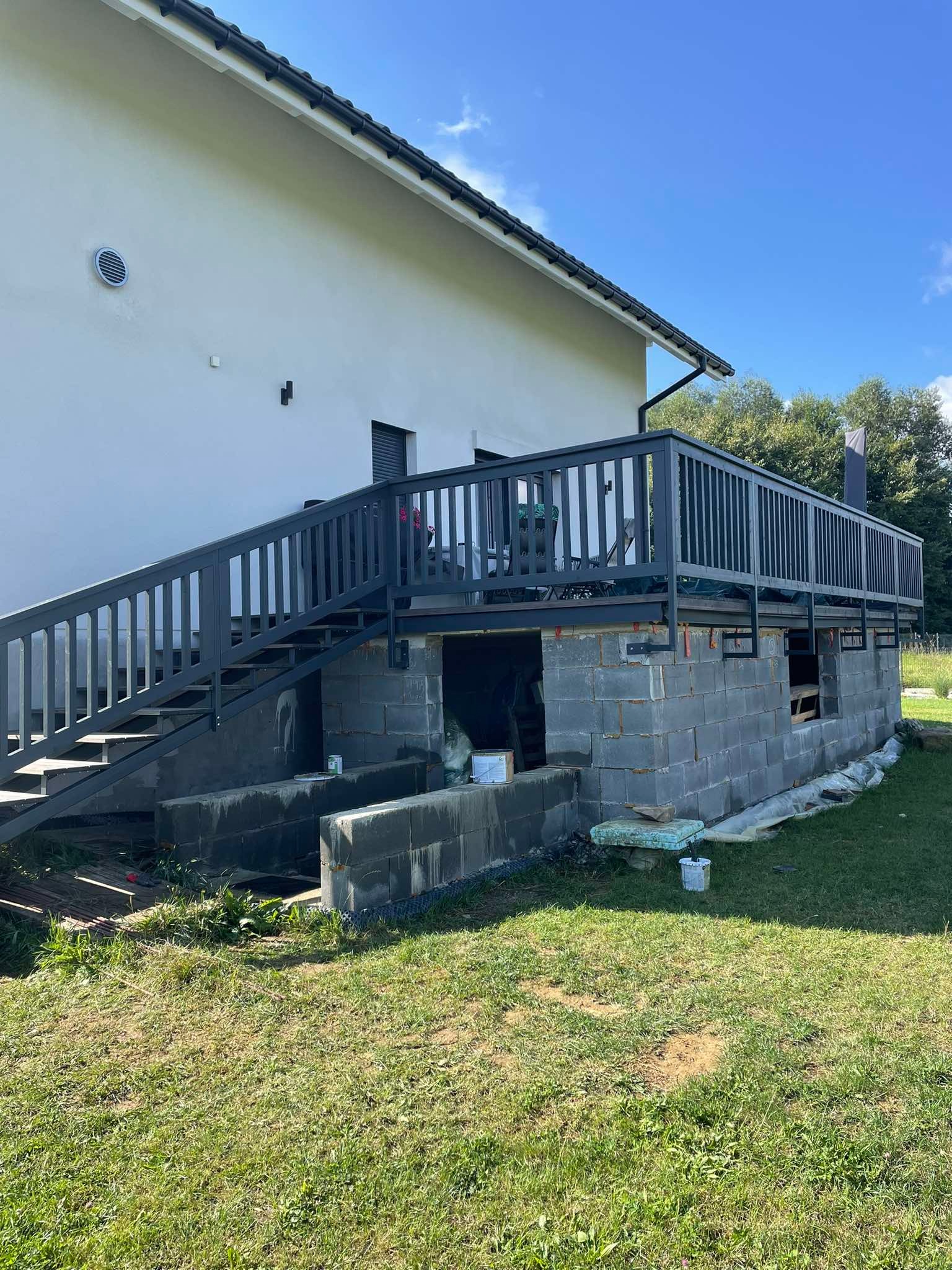 Wznoszenie tarasu z szarą balustradą i schodami, widok na surową konstrukcję z bloczków betonowych, farba i materiały budowlane w pobliżu, dom w tle.