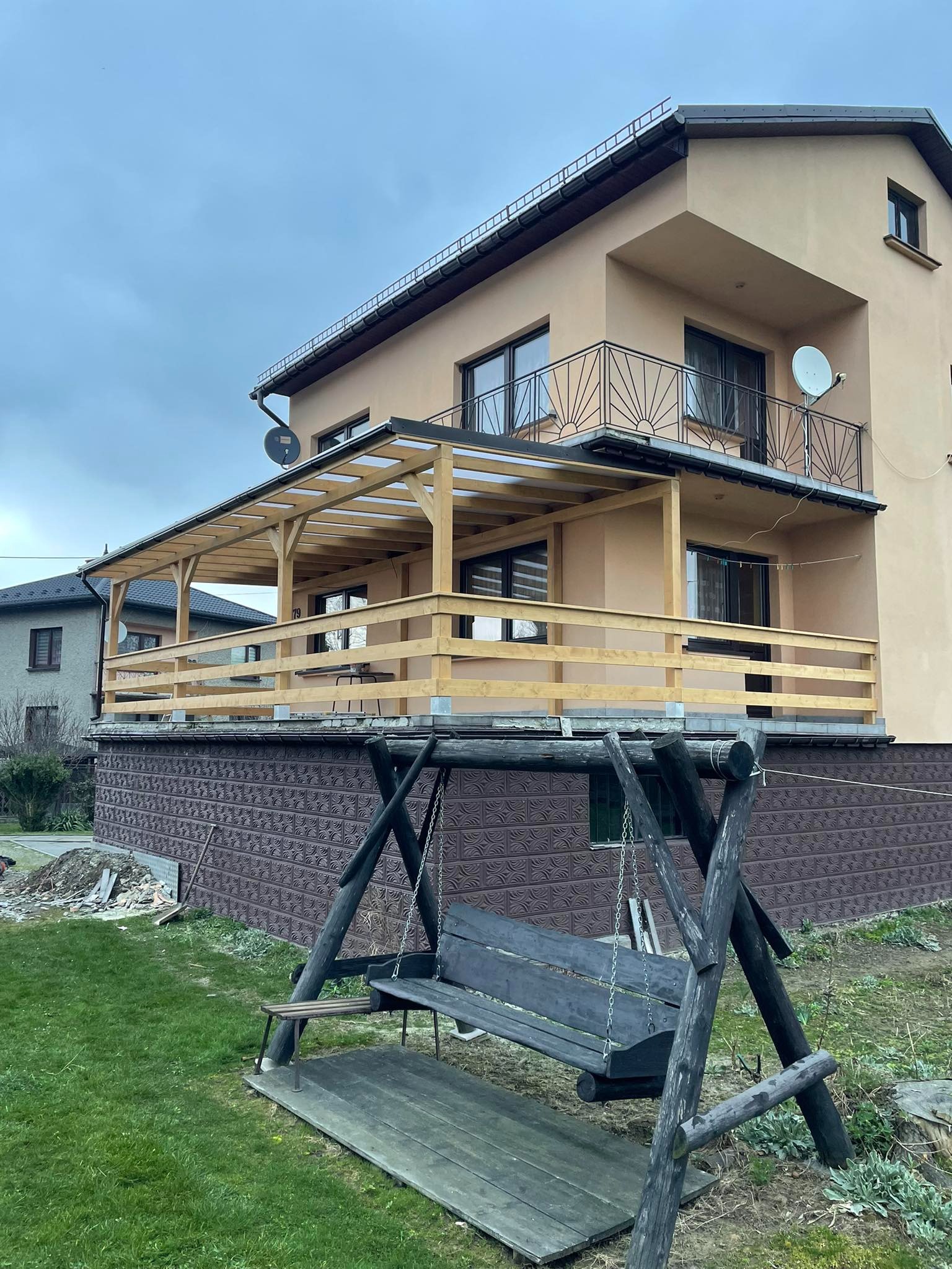 Dwupiętrowy dom z beżową elewacją, balkonem z czarną balustradą i drewnianym tarasem na podwyższeniu z widoczną konstrukcją zadaszenia, w tle sąsiedni dom i pochmurne niebo, na pierwszym planie...