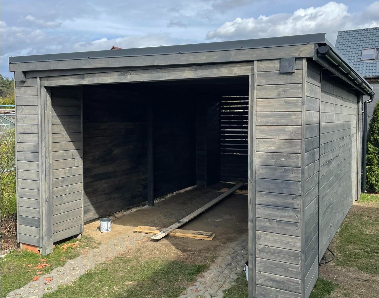 Nowoczesna, otwarta konstrukcja z szarego drewna z płaskim dachem i widocznymi śladami impregnacji, częściowo wykończona, z widocznymi deskami i wiadrem farby w środku.