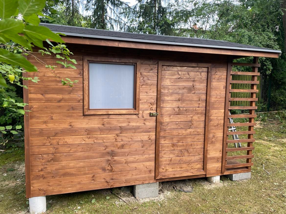 Drewniana altana ogrodowa z oknem z pleksi i kratką, podniesiona na betonowych bloczkach, otoczona zielenią.