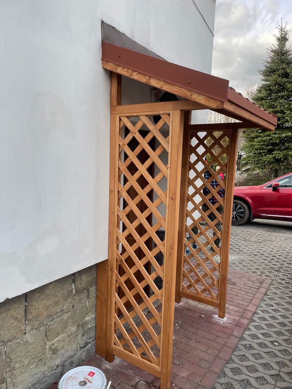 Drewniana konstrukcja z kratownicą, pełniąca funkcję zadaszenia wejściowego, przylegająca do jasnej ściany budynku z kamienną podmurówką, widoczny fragment czerwonego samochodu i choinki w tle.