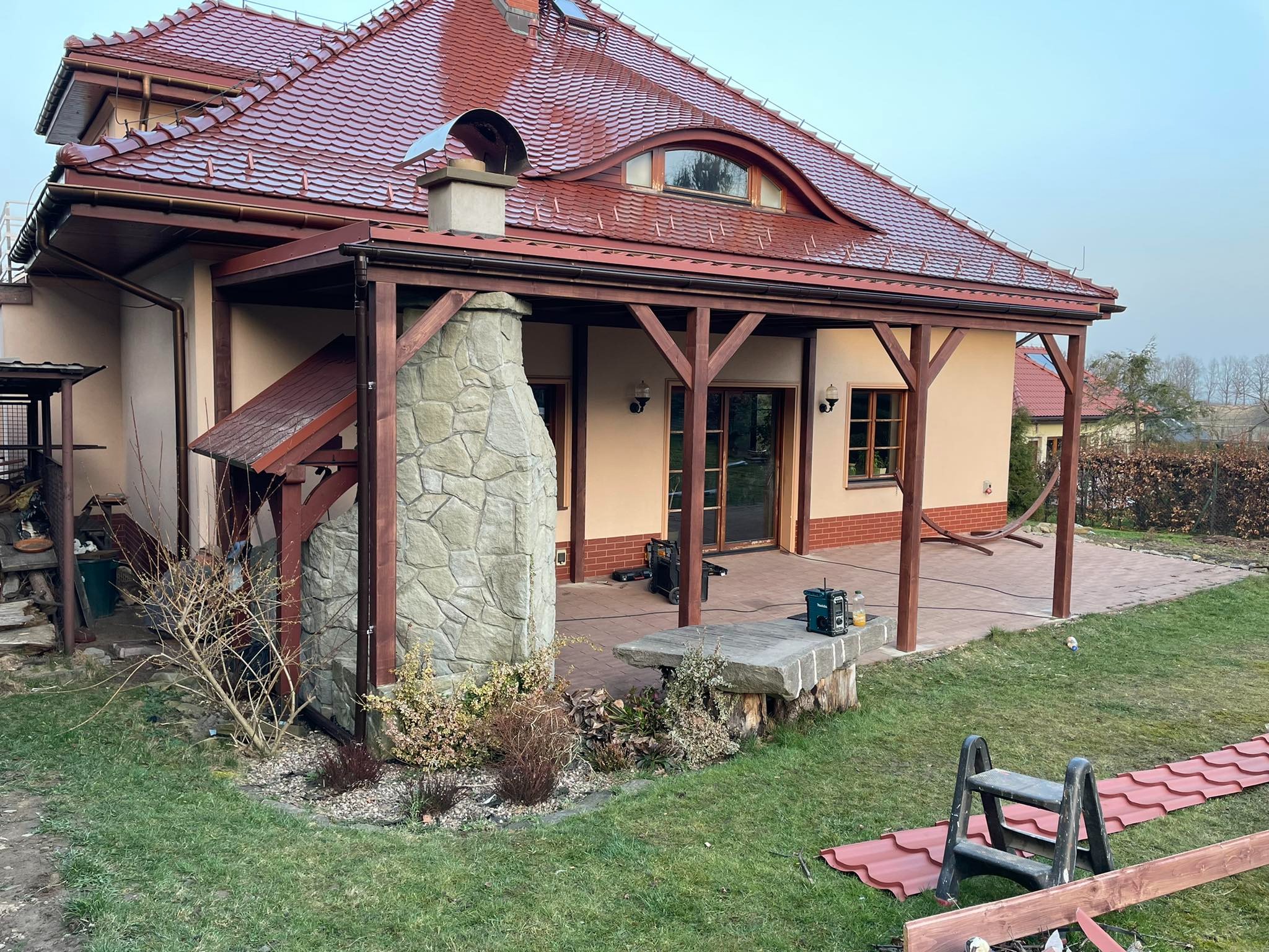 Zadaszony taras z drewnianą konstrukcją i kamiennym kominkiem przylegający do domu z czerwoną dachówką, widoczny fragment ogrodu i drabina ze zdjętymi dachówkami.