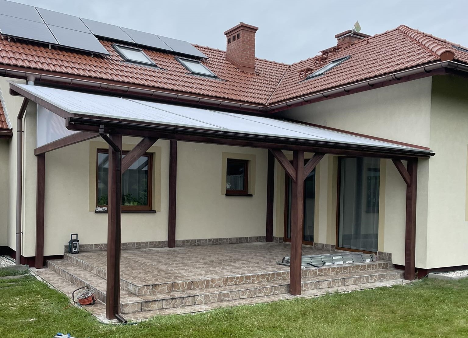 Zadaszony taras z brązową drewnianą konstrukcją przy beżowym domu z brązową dachówką i panelami słonecznymi, widoczne schody z płytek i drabina.