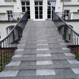 FAST BILDING VASYL VOLOSHYN - Zewnętrzne schody wejściowe do białego budynku, wyłożone szarymi płytkami w dwóch odcieniach, z czarną metalową balustradą.