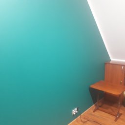 FAST BILDING VASYL VOLOSHYN - Turkusowa ściana w pokoju na poddaszu, z drewnianym biurkiem i szafką w rogu, widoczne gniazdko elektryczne przy podłodze.