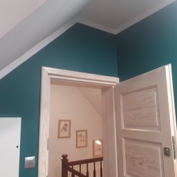 FAST BILDING VASYL VOLOSHYN - Szmaragdowa ściana z białą listwą przypodłogową i ościeżnicą z surowego drewna, widok na klatkę schodową z trzema obrazkami w złotych ramkach.