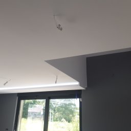 FAST BILDING VASYL VOLOSHYN - Nowoczesne wnętrze z szarą ścianą, białym sufitem z podświetleniem LED i dużym oknem z widokiem na zieleń, widoczne kable od oświetlenia sufitowego w trakcie montażu.