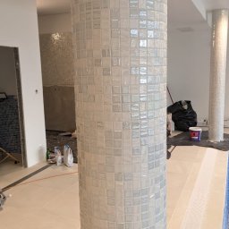 FAST BILDING VASYL VOLOSHYN - Biała mozaika kwadratowa na kolumnie w trakcie montażu, widoczne fugi i fragmenty innych elementów wykończeniowych w tle, w tym podłoga z odpływem liniowym.