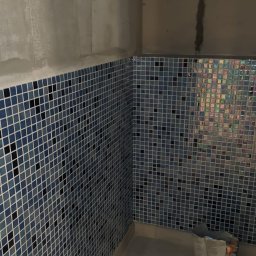 FAST BILDING VASYL VOLOSHYN - Narożnik łazienki w trakcie wykańczania: ściany wyłożone niebieską mozaiką, surowy beton na górze, widoczne ślady zaprawy, na podłodze wiadro z resztkami materiałów budowlanych.