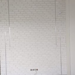 FAST BILDING VASYL VOLOSHYN - Ściana wykończona białą cegłą dekoracyjną z widocznymi przewodami elektrycznymi w otworach gniazdek oraz profilami aluminiowymi zamontowanymi pionowo na ścianie.