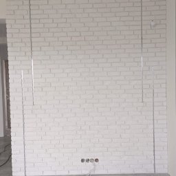 FAST BILDING VASYL VOLOSHYN - Ściana z białej cegły dekoracyjnej z widocznymi metalowymi listwami pionowymi, przygotowana pod montaż instalacji elektrycznej, na podłodze szare płytki.