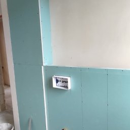 FAST BILDING VASYL VOLOSHYN - Montaż instalacji elektrycznej i hydraulicznej w ścianie z zielonej płyty gipsowo-kartonowej, widoczne kable, rury, puszka elektryczna, wiertarka i narzędzia na podłodze.