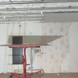 FAST BILDING VASYL VOLOSHYN - Montaż sufitu podwieszanego: podkonstrukcja z profili stalowych i częściowo zamontowana płyta na podnośniku w pomieszczeniu z widocznymi fragmentami starej tapety i resztkami farby na ścianach.