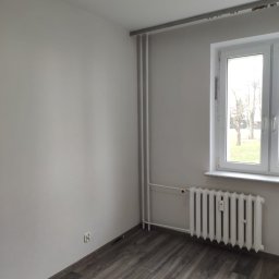 FAST BILDING VASYL VOLOSHYN - Pokój po remoncie w Stargardzie: świeżo pomalowane szare ściany, ułożone panele podłogowe imitujące drewno, biały grzejnik pod oknem z widokiem na drzewa bez liści.