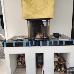 FAST BILDING VASYL VOLOSHYN - Montaż obudowy kominka z czarnym blatem, zabezpieczonym niebieską taśmą, złota górna część, wnęki z drewnem opałowym i brykietem, widoczne narzędzia i odblask osoby robiącej zdjęcie.