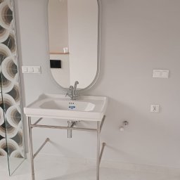 FAST BILDING VASYL VOLOSHYN - Biała umywalka z chromowanym stelażem i baterią, owalne lustro w srebrnej ramie, szare ściany, widoczne włączniki i gniazdko elektryczne, fragment kabiny prysznicowej z wzorzystymi płytkami w tle.