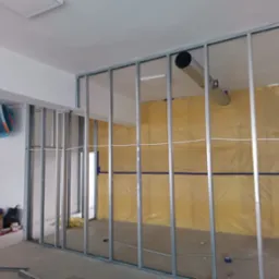 Metalowy szkielet ścian działowych, ocieplony żółtą wełną mineralną, widoczny w trakcie montażu w nowym pomieszczeniu, z fragmentem instalacji wentylacyjnej pod sufitem i narzędziami na podłodze.