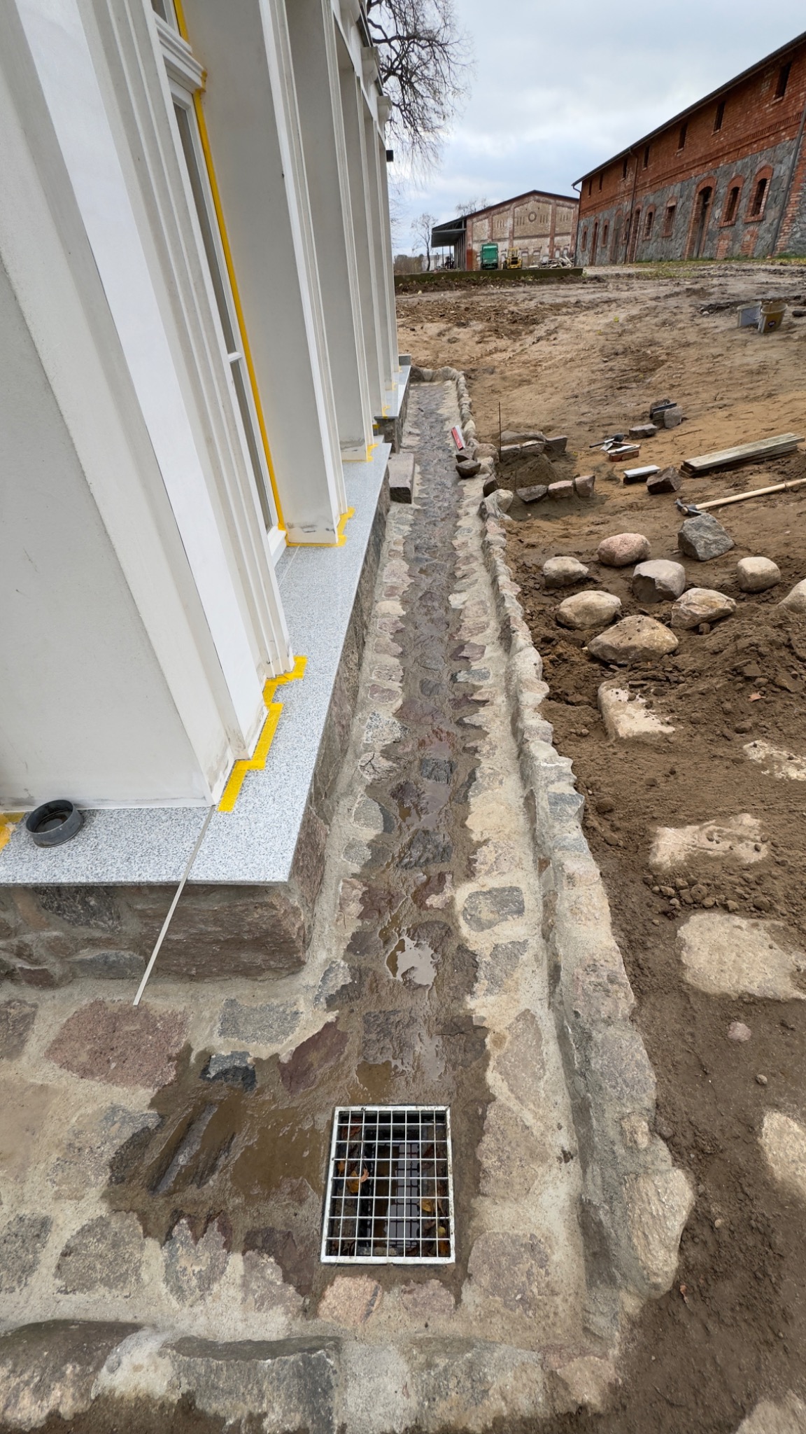 Kamienny odpływ liniowy z kratką i wodą, przylegający do elewacji budynku z oknami zabezpieczonymi żółtą taśmą malarską, w tle budynki gospodarcze i ziemia.