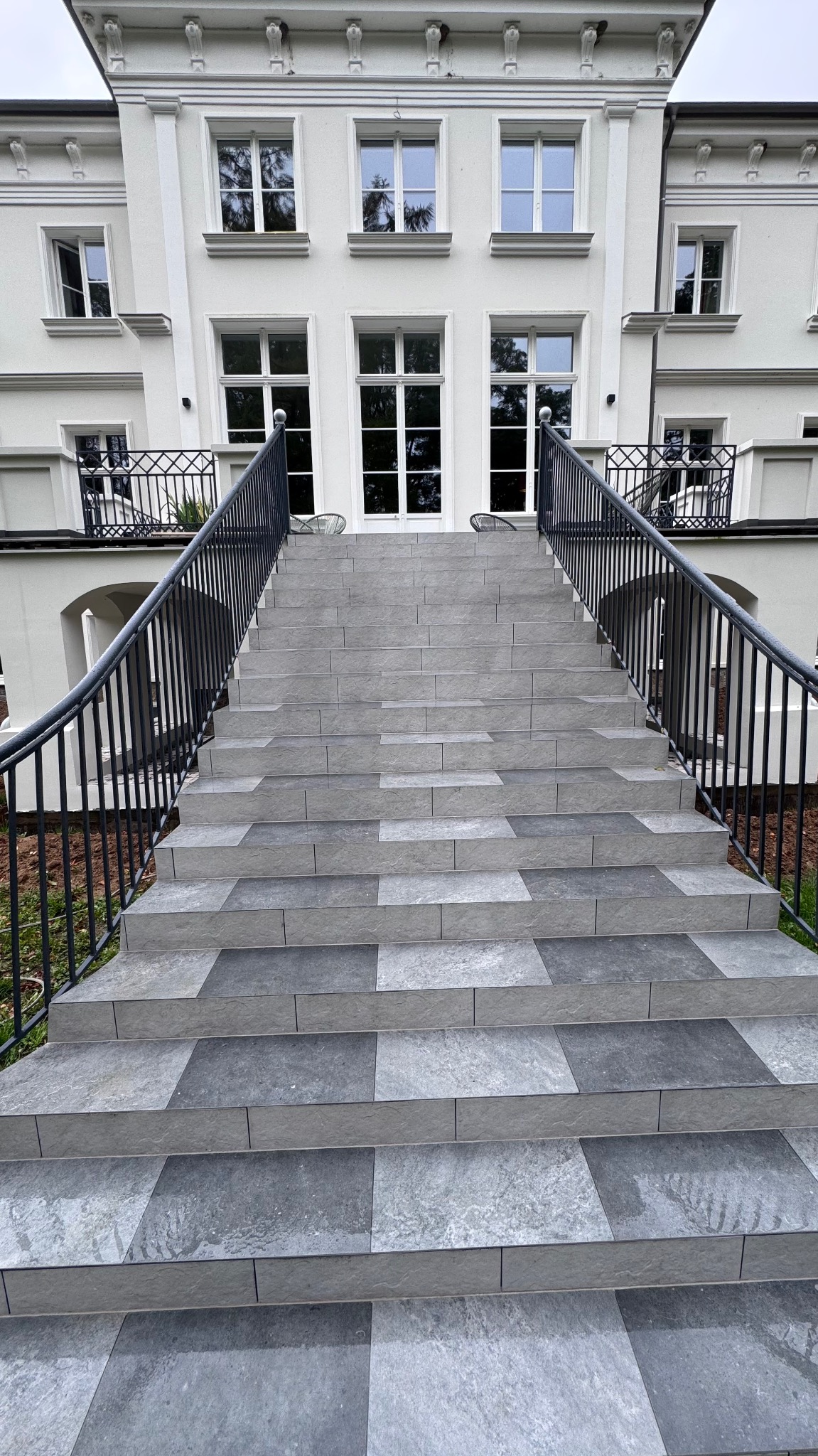 Zewnętrzne schody wejściowe do białego budynku, wyłożone szarymi płytkami w dwóch odcieniach, z czarną metalową balustradą.
