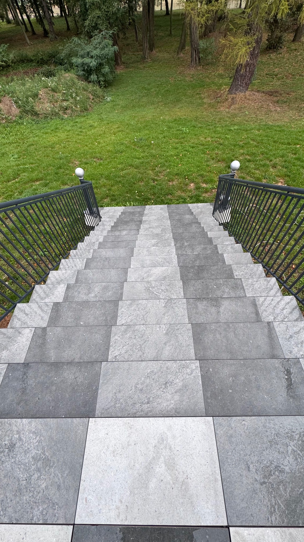 Zewnętrzne schody wyłożone szarymi płytami o zróżnicowanej fakturze, prowadzące do zielonego trawnika i drzew, zabezpieczone czarną metalową balustradą z białymi kulami na końcach.
