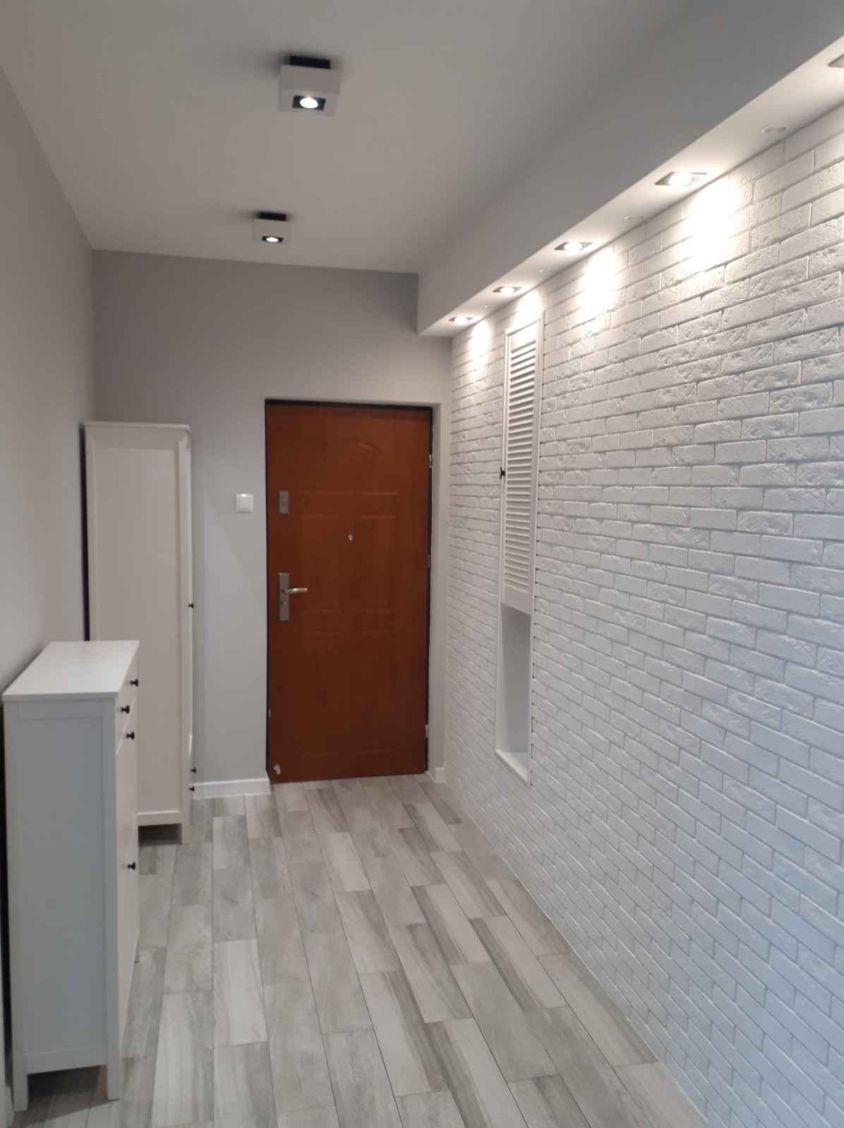 Przedpokój po remoncie: biała ściana z cegieł, drewniane panele podłogowe w odcieniach szarości, brązowe drzwi wejściowe, biała szafka z szufladami i wbudowane oświetlenie sufitowe.