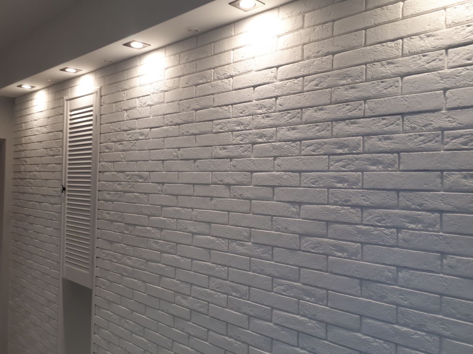 Ściana z białej cegły dekoracyjnej z widocznymi fugami, oświetlona punktowymi lampami sufitowymi, z białymi, żaluzjowymi drzwiczkami rewizyjnymi po lewej stronie.