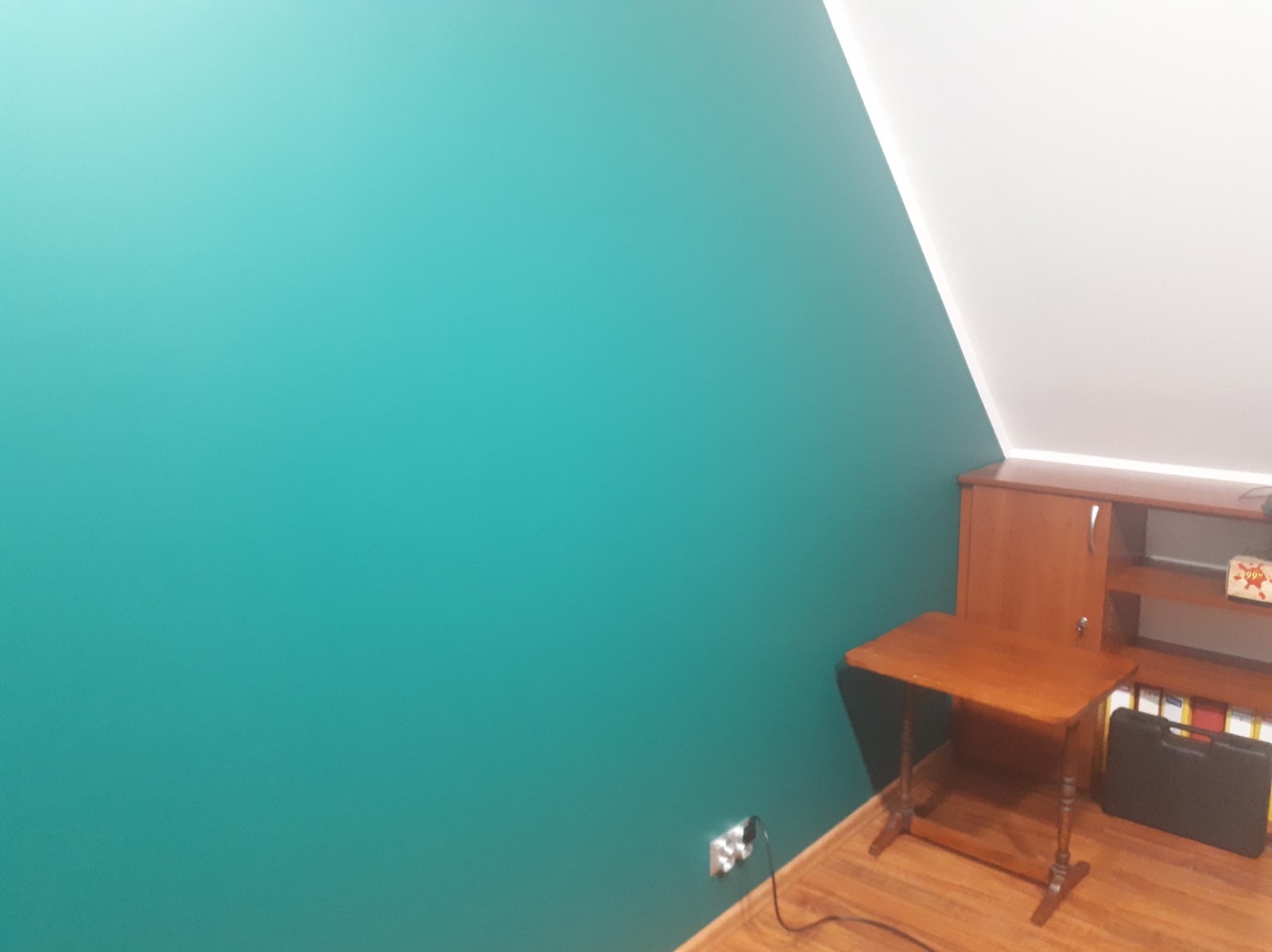 Turkusowa ściana w pokoju na poddaszu, z drewnianym biurkiem i szafką w rogu, widoczne gniazdko elektryczne przy podłodze.