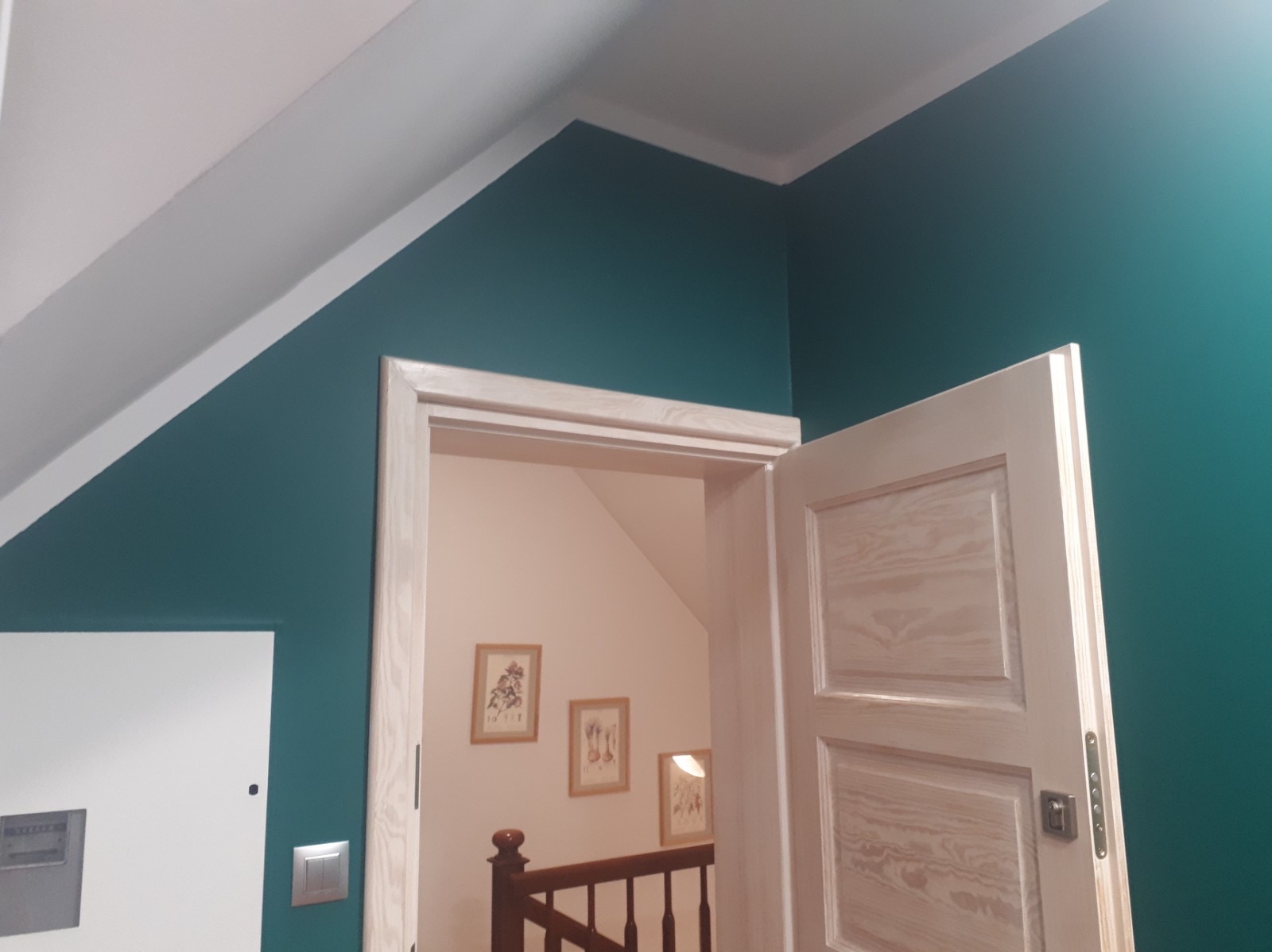 Szmaragdowa ściana z białą listwą przypodłogową i ościeżnicą z surowego drewna, widok na klatkę schodową z trzema obrazkami w złotych ramkach.