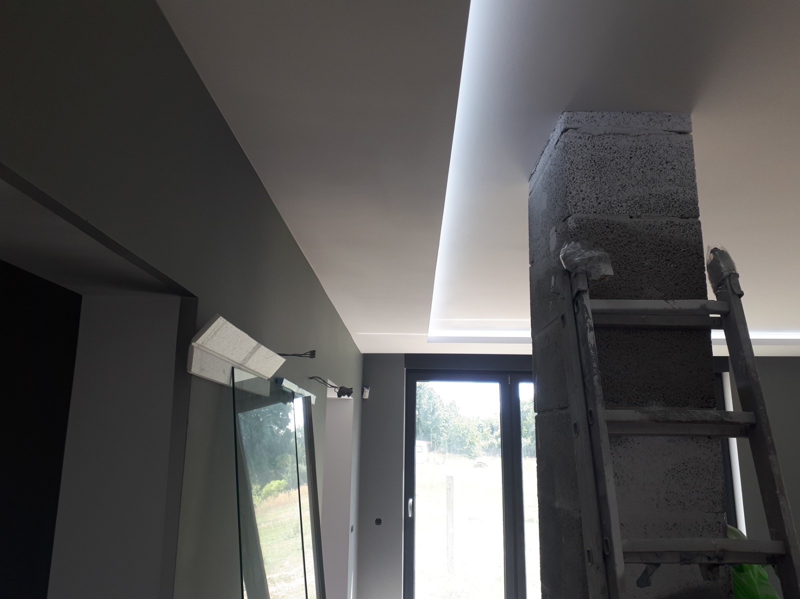 Wnętrze w trakcie prac wykończeniowych: szara ściana, sufit z oświetleniem LED, kolumna z betonu komórkowego z drabiną, widok na okno.