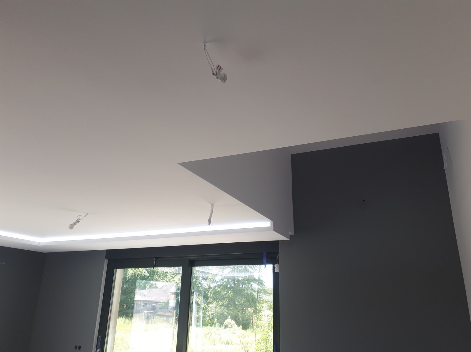 Nowoczesne wnętrze z szarą ścianą, białym sufitem z podświetleniem LED i dużym oknem z widokiem na zieleń, widoczne kable od oświetlenia sufitowego w trakcie montażu.