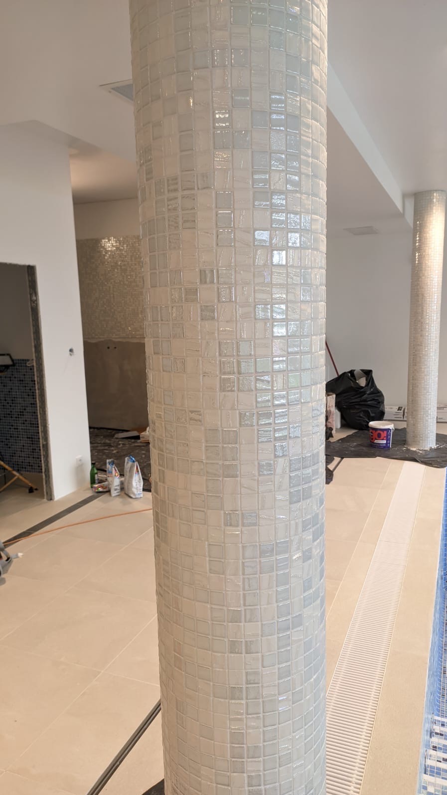 Biała mozaika kwadratowa na kolumnie w trakcie montażu, widoczne fugi i fragmenty innych elementów wykończeniowych w tle, w tym podłoga z odpływem liniowym.