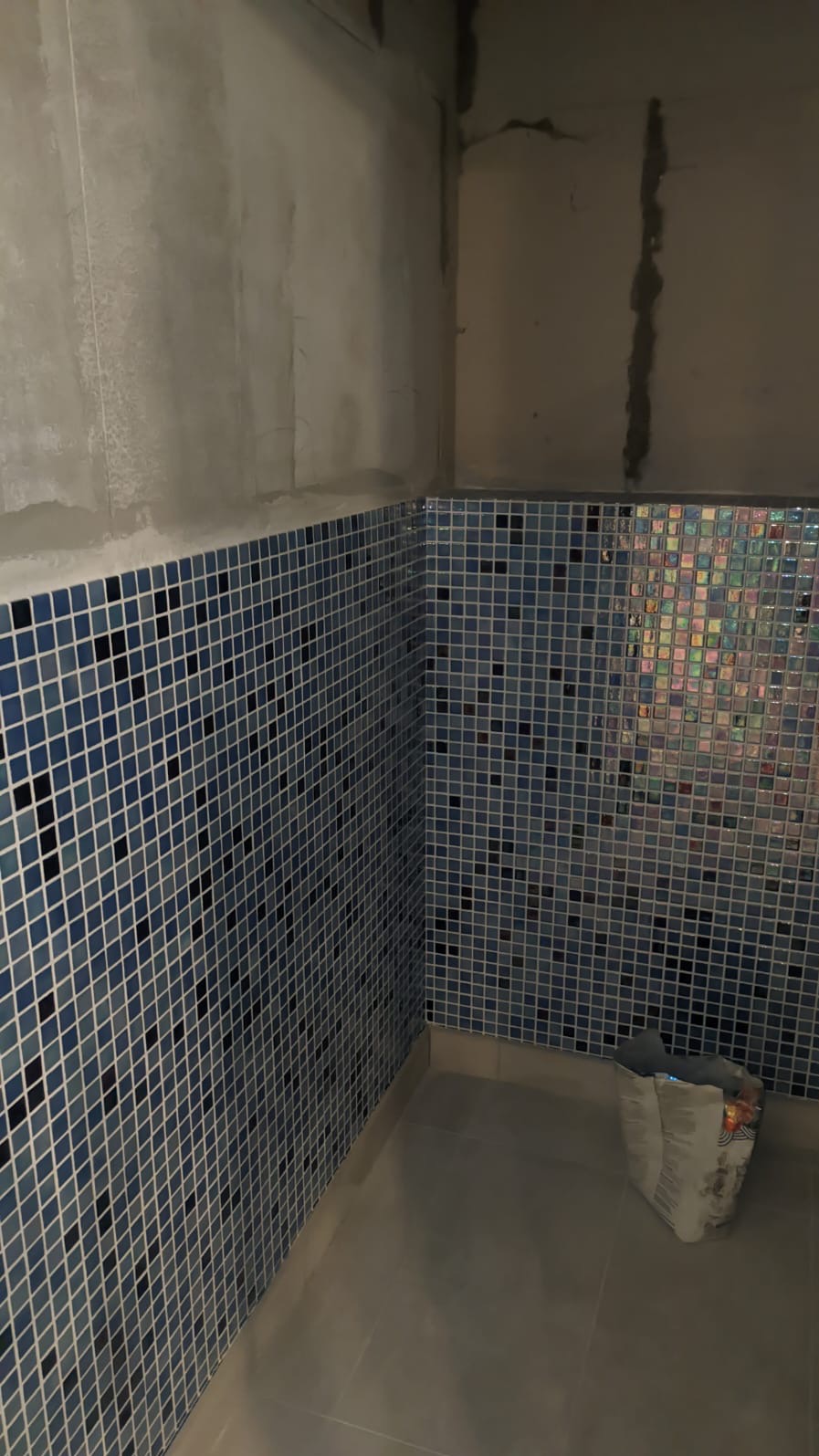 Narożnik łazienki w trakcie wykańczania: ściany wyłożone niebieską mozaiką, surowy beton na górze, widoczne ślady zaprawy, na podłodze wiadro z resztkami materiałów budowlanych.