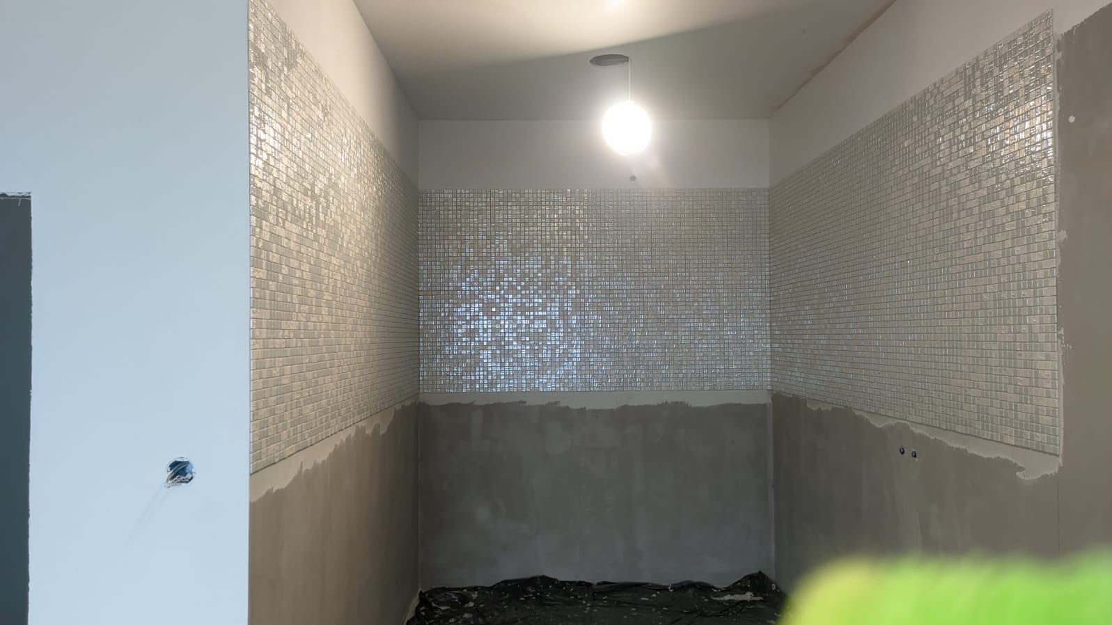 Montaż błyszczącej mozaiki na ścianach w łazience, widoczne niedociągnięcia w fugowaniu i przygotowaniu podłoża, surowe wykończenie w trakcie prac.