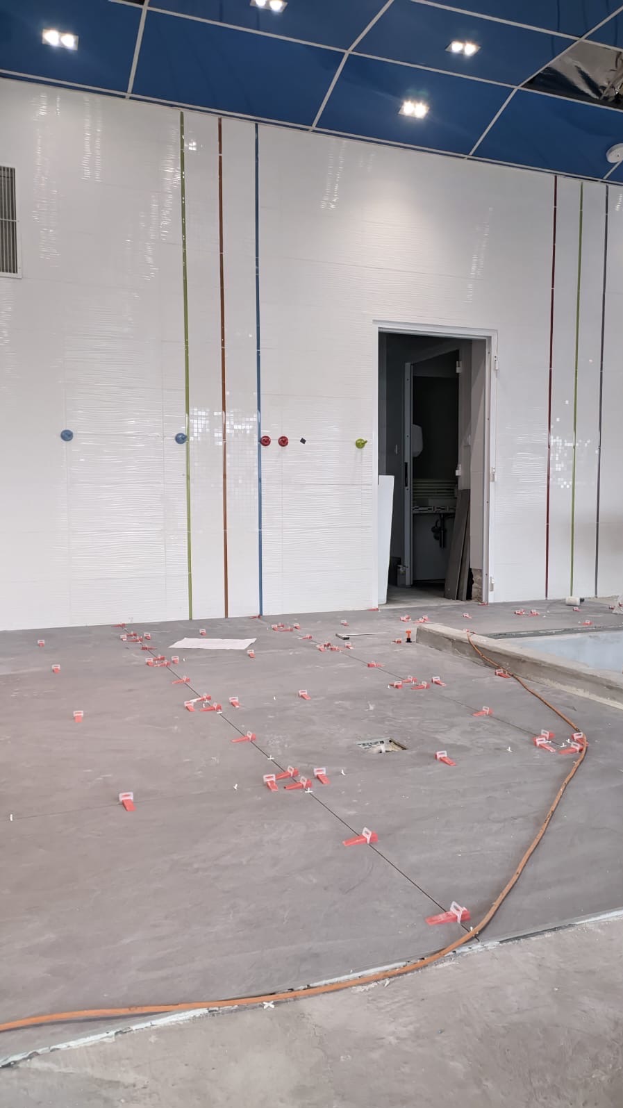 Wnętrze w trakcie prac wykończeniowych: białe płytki ścienne z delikatnym wzorem fal, pionowe kolorowe paski na ścianie, poziomowanie podłogi z użyciem krzyżyków dystansowych i sznurka, widoczne...