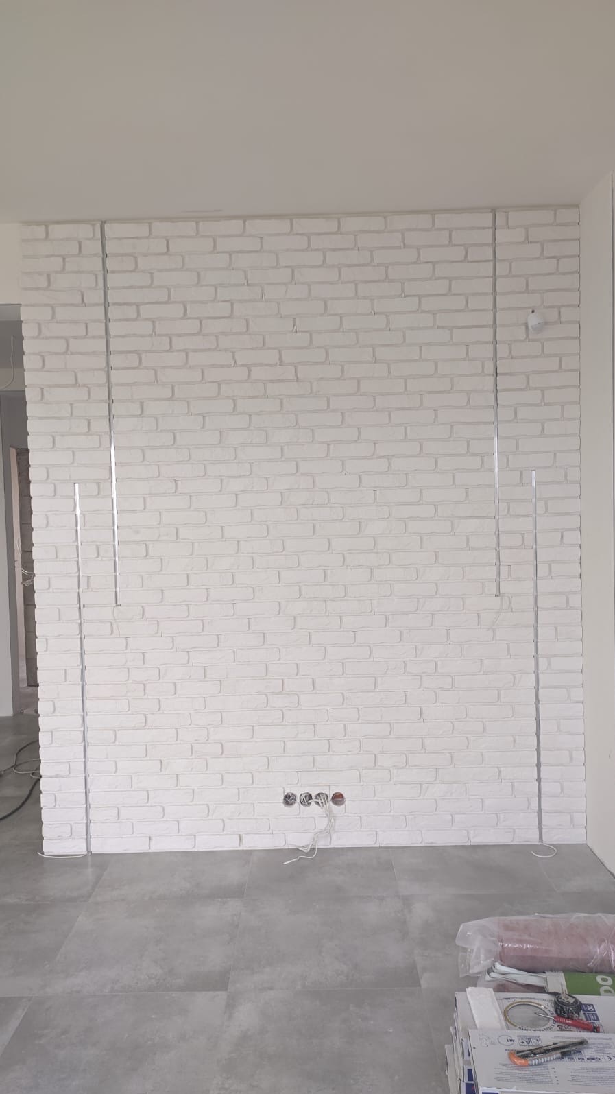Ściana z białej cegły dekoracyjnej z widocznymi metalowymi listwami pionowymi, przygotowana pod montaż instalacji elektrycznej, na podłodze szare płytki.