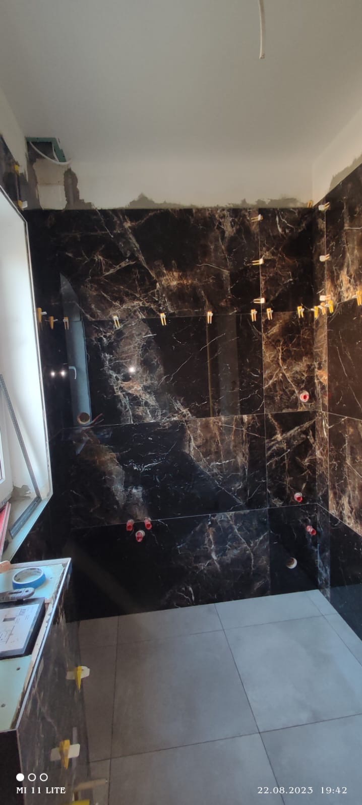 Proces układania płytek imitujących marmur na ścianie łazienki, widoczne plastikowe kliny poziomujące i otwory na instalacje, szare płytki podłogowe, surowy stan wykończenia sufitu.