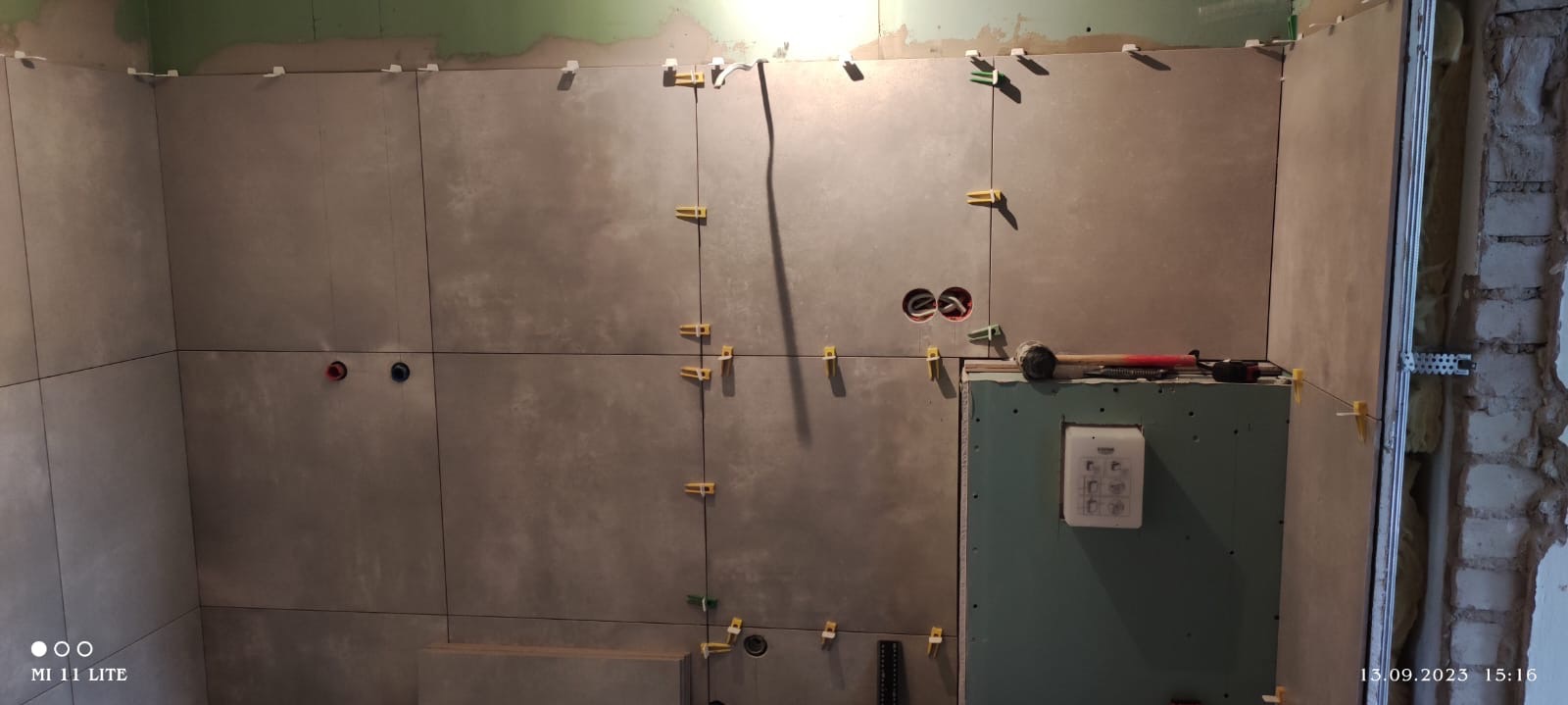 Szare płytki ceramiczne układane na ścianie z widocznymi otworami na instalacje i żółtymi klipsami poziomującymi, młotek gumowy leżący na płycie kartonowo-gipsowej obok włącznika podtynkowego.