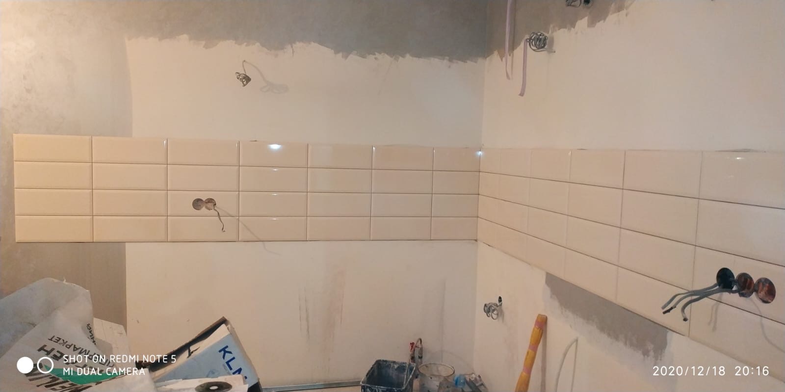 Ściana w trakcie układania beżowych, prostokątnych płytek, widoczne puszki elektryczne z wystającymi kablami, surowy tynk na górze ściany, narzędzia i materiały budowlane w dolnej części kadru.