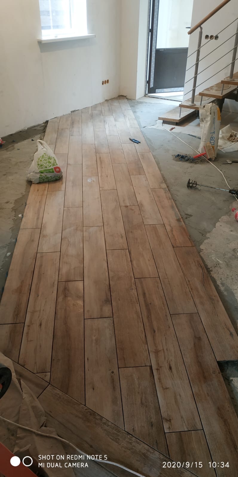 Układanie podłogi z drewnopodobnych płytek w trakcie remontu, widoczny worek z klejem, mikser do zaprawy, poziomica i schody w tle.