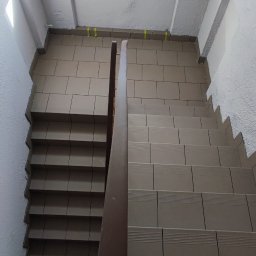 SaVo - Klatka schodowa w trakcie wykańczania, widok z góry na brązowe płytki na schodach i żółtą taśmę maskującą przy suficie, drewniana poręcz i fragment stopy w klapku.