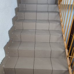 SaVo - Układanie szarych płytek na schodach w klatce schodowej, widoczne plastikowe krzyżyki dystansowe, butelka wody i pracownik w tle.