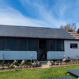 SaVo - Dom z blachy falistej na elewacji i dachu, z widocznymi dziećmi w drzwiach, ogródkiem skalnym i fragmentami blachy na trawniku.