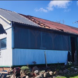 SaVo - Dom z częściowo zdjętym, rdzawym dachem z blachy falistej, fragment dachu pokryty nową szarą blachą, widoczne krokwie, narzędzia budowlane leżące obok domu, niebieskie niebo z chmurami w tle.