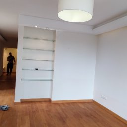 SaVo - Pusta, jasna przestrzeń pokoju z drewnianą podłogą, białymi ścianami, wbudowanymi szklanymi półkami i widoczną osobą w przejściu do kolejnego pomieszczenia.