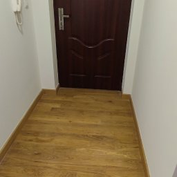 SaVo - Widok na świeżo położoną podłogę z paneli imitujących drewno, prowadzącą do ciemnobrązowych drzwi wejściowych w białym korytarzu. Na ścianie widoczny biały domofon i skrzynka elektryczna.